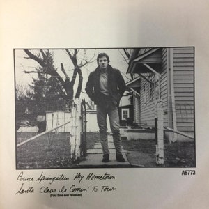 Puede incluir: Foto en blanco y negro de Bruce Springsteen de pie frente a una casa. La foto está firmada "Bruce Springsteen My Hometown Santa Claus Is Comin' To Town (First time ever released)" y "A6773".
