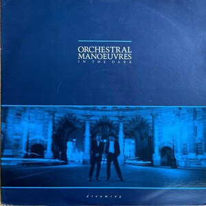 Może przedstawiać: Niebieska okładka płyty winylowej z tekstem "Orchestral Manoeuvres In The Dark" i "dreaming" w kolorze białym. Okładka przedstawia rozmyte zdjęcie dwóch mężczyzn w garniturach stojących przed dużym łukiem.
