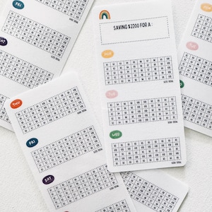 May include: White rectangular savings tracker cards with labels for each day of the week. Each card has a grid with numbers and the text "SAVING £2200 FOR A:" with a rainbow graphic. The cards are designed for financial planning.