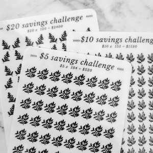 May include: Three white sheets of paper with black text and black leaf designs. The top sheet says "&pound;20 savings challenge" with the equation "&pound;20 x 120 = &pound;2400". The middle sheet says "&pound;10 savings challenge" with the equation "&pound;10 x 153 = &pound;1530". The bottom sheet says "&pound;5 savings challenge" with the equation "&pound;5 x 104 = &pound;520".
