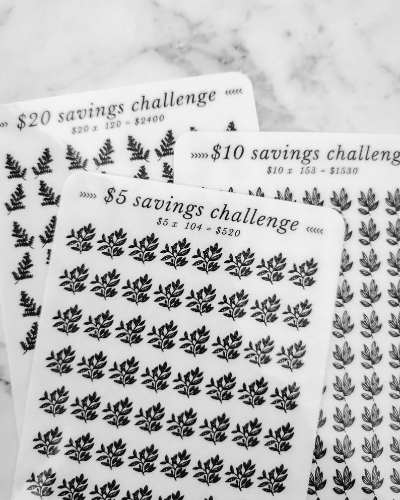 May include: Three white sheets of paper with black text and black leaf designs. The top sheet says "&pound;20 savings challenge" with the equation "&pound;20 x 120 = &pound;2400". The middle sheet says "&pound;10 savings challenge" with the equation "&pound;10 x 153 = &pound;1530". The bottom sheet says "&pound;5 savings challenge" with the equation "&pound;5 x 104 = &pound;520".