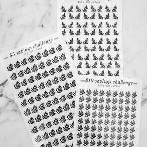 May include: Three sheets of stickers with black leaf designs. Each sheet has a different savings challenge printed on it: &pound;5 savings challenge, &pound;10 savings challenge, and &pound;20 savings challenge. The sheets are on a white marble surface.
