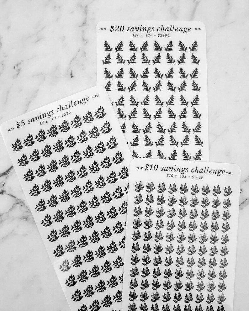 May include: Three sheets of stickers with black leaf designs. Each sheet has a different savings challenge printed on it: &pound;5 savings challenge, &pound;10 savings challenge, and &pound;20 savings challenge. The sheets are on a white marble surface.