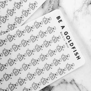 May include: Two white cards with rounded corners, featuring black line drawings of goldfish in bags. One card says "BE A GOLDFISH" with text below. The other card has "FISH" at the top. The background is a marble surface.