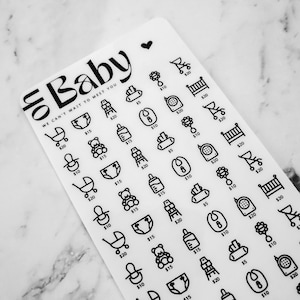 May include: A white sticker sheet with black baby-themed icons and prices. The icons include a baby bottle, a rattle, a crib, a diaper, a teddy bear, a stroller, a high chair, and a baby monitor. The text "Baby" is at the top of the sheet, with the words "We can't wait to meet you" below it.