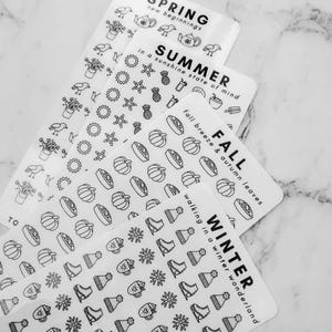 May include: Four sheets of white sticker paper with black outlines of various seasonal icons. The top sheet says "SPRING new beginnings" and features icons like a teapot, birds, and plants. The second sheet says "SUMMER in a sunshine state of mind" and features icons like pineapples, sunglasses, and seashells. The third sheet says "FALL fall breeze & autumn leaves" and features icons like pumpkins, pies, and leaves. The bottom sheet says "WINTER walking in a winter wonderland" and features icons like snowflakes, ice skates, and mugs.