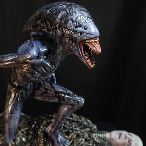 Könnte beinhalten: Eine detaillierte Skulptur einer Alien-Kreatur, die über einer menschlichen Figur steht. Der Alien ist dunkelblau mit einem strukturierten Kopf und offenem Mund. Die menschliche Figur ist blass mit einem metallischen Arm. Die Basis ist grau mit dem Text "FINAL PRODUCT".