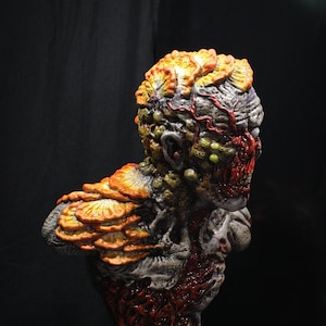 Pode incluir: Um busto detalhado de um Clicker do jogo The Last of Us. O Clicker é um humano infectado por fungos com uma cabeça grande e bulbosa coberta por crescimentos fúngicos de cor laranja e amarela. A pele do Clicker é vermelha e carnuda, com veias e tendões visíveis. O busto está montado em uma base circular com as palavras "The Last of Us" impressas nela.