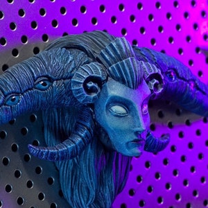 Puede incluir: Una figura esculpida de color azul y negro de una criatura con cuernos y múltiples ojos. La figura está montada en una pared de tablero de clavijas con una luz morada que brilla sobre ella. El texto "PINOCCHIO DE GUILLERMO DEL TORO" es visible sobre la figura.