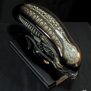 Puede incluir: Una escultura detallada de una cabeza alienígena, con un diseño elegante y alargado y elementos mecánicos y orgánicos intrincados. La cabeza es principalmente oscura con acentos metálicos y se asienta sobre una base rectangular negra. Producto final.