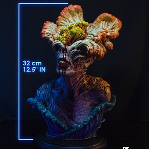 May include: A detailed bust of a creature from "The Last of Us" video game, featuring a grotesque, fungal-covered head and chest. The sculpture is approximately 12.5 inches (32 cm) tall, with a dark base and intricate paintwork. The bust has a dark background.
