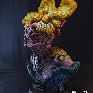 May include: A detailed bust of a Clicker from The Last of Us. The sculpture features a grotesque, mutated human head with fungal growth in shades of yellow, orange, and green. The bust is mounted on a black base, with the game's logo visible.