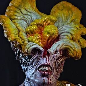 May include: A detailed sculpture of a Clicker from The Last of Us. The figure features a grotesque, mutated face with exposed teeth, blood, and fungal growth in shades of yellow, orange, and gray. The sculpture has the text "The Last of Us" and "Crisis Estudio".