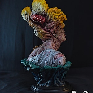 May include: A detailed bust of a Clicker from The Last of Us, featuring a grotesque, fungal head in shades of yellow, red, and brown. The figure has a decaying appearance, with a textured, zombie-like skin and a tattered shirt. The base is black.