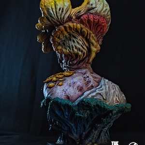 May include: A detailed bust of a Clicker from The Last of Us, featuring a grotesque, fungal head in shades of yellow, orange, and red. The bust has a decaying, textured appearance with a purple and white torso, and is set on a black base.