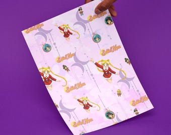 Puede incluir: Una hoja rectangular de papel de regalo rosa claro con un patrón repetitivo de personajes de Sailor Moon en trajes rojos y blancos, logotipos dorados de Sailor Moon, lunas crecientes, estrellas y adornos circulares con retratos.