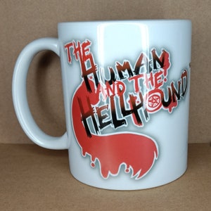 Puede incluir: Taza de cerámica blanca con un diseño gráfico rojo y negro que dice "The Human and the Hellhound".