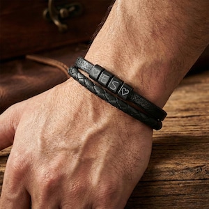 Pode incluir: Duas pulseiras de couro preto são usadas no pulso. Uma pulseira é trançada e a outra tem três contas pretas com as letras "V", "E" e "S" e um símbolo de coração. As pulseiras estão no pulso de uma pessoa.