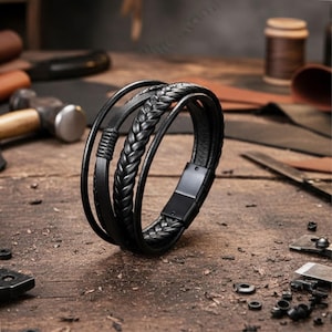 Può includere: Bracciale in pelle nera con più fili, tra cui una sezione intrecciata. Il bracciale presenta una chiusura magnetica ed è esposto su una superficie di legno, evidenziando il suo aspetto artigianale. Un accessorio elegante.
