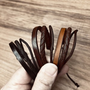 May include: A set of five leather bracelets in brown and black colours. The bracelets are braided and have a simple design.