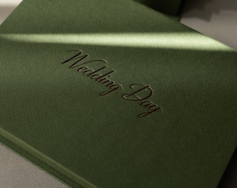 May include: Olive green wedding album with the embossed text "Wedding Day" in a script font. The album is rectangular and appears to be made of a textured material. Several albums are stacked, with shadows across the covers.