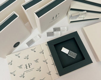 May include: Cream-colored boxes with dark green interiors and the text "HANNAH PHIPPS PHOTOGRAPHY" are arranged on a white surface. Some boxes are open, revealing clear USB drives. One box features a hummingbird pattern.
