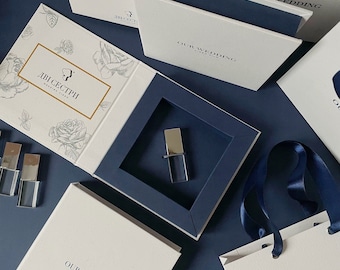 May include: A white box with a blue interior and a clear USB drive inside. The box is labeled "OUR WEDDING VIDEO FILM" and has a floral design. The box is sitting on a blue surface.