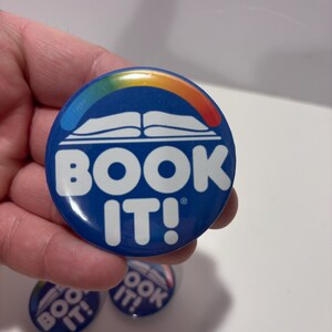 K&ouml;nnte beinhalten: Ein runder blauer Ansteckbutton mit einer wei&szlig;en Grafik eines offenen Buches, das von einem Regenbogenbogen gekr&ouml;nt wird. Der Text "BOOK IT!" ist in gro&szlig;en wei&szlig;en Gro&szlig;buchstaben unter der Buchgrafik gedruckt. Der Button hat eine gl&auml;nzende Oberfl&auml;che und ein kleines Markenzeichen-Symbol.