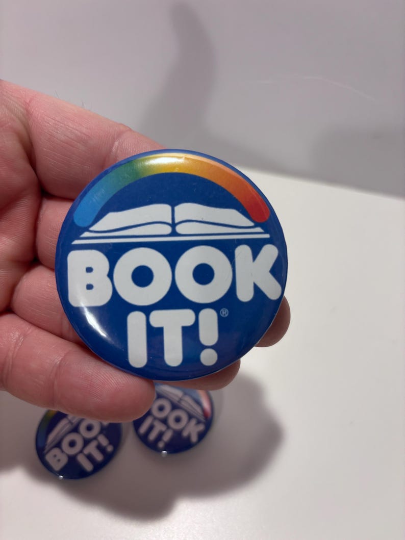 K&ouml;nnte beinhalten: Ein runder blauer Ansteckbutton mit einer wei&szlig;en Grafik eines offenen Buches, das von einem Regenbogenbogen gekr&ouml;nt wird. Der Text "BOOK IT!" ist in gro&szlig;en wei&szlig;en Gro&szlig;buchstaben unter der Buchgrafik gedruckt. Der Button hat eine gl&auml;nzende Oberfl&auml;che und ein kleines Markenzeichen-Symbol.