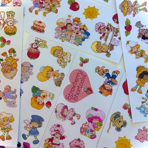 Può includere: Una collezione di adesivi Strawberry Shortcake su fogli bianchi con vari personaggi, torte, fragole e soli. Una grafica a forma di cuore rosa al centro mostra il testo Strawberry Shortcake in caratteri rosa scuro.