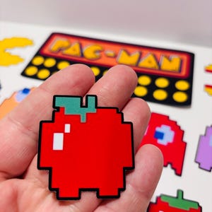 以下が含まれることがあります： 緑の茎と白いハイライトが付いたピクセル化された赤いリンゴのステッカー。このデザインは、黒い輪郭を持つブロック状のレトロなビデオゲームの美学を特徴としています。ステッカーは、パックマンを含む他のゲームをテーマにしたステッカーの背景に対して表示されています。