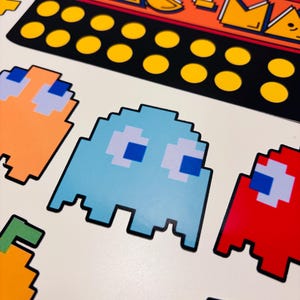 以下が含まれることがあります： ピクセル化されたパックマンのキャラクターとゲーム要素が描かれたステッカーシート。赤、オレンジ、水色のゴースト、黄色のドット、そしてオレンジと黄色の文字でPAC-MANと書かれた黒い長方形のロゴが含まれています。