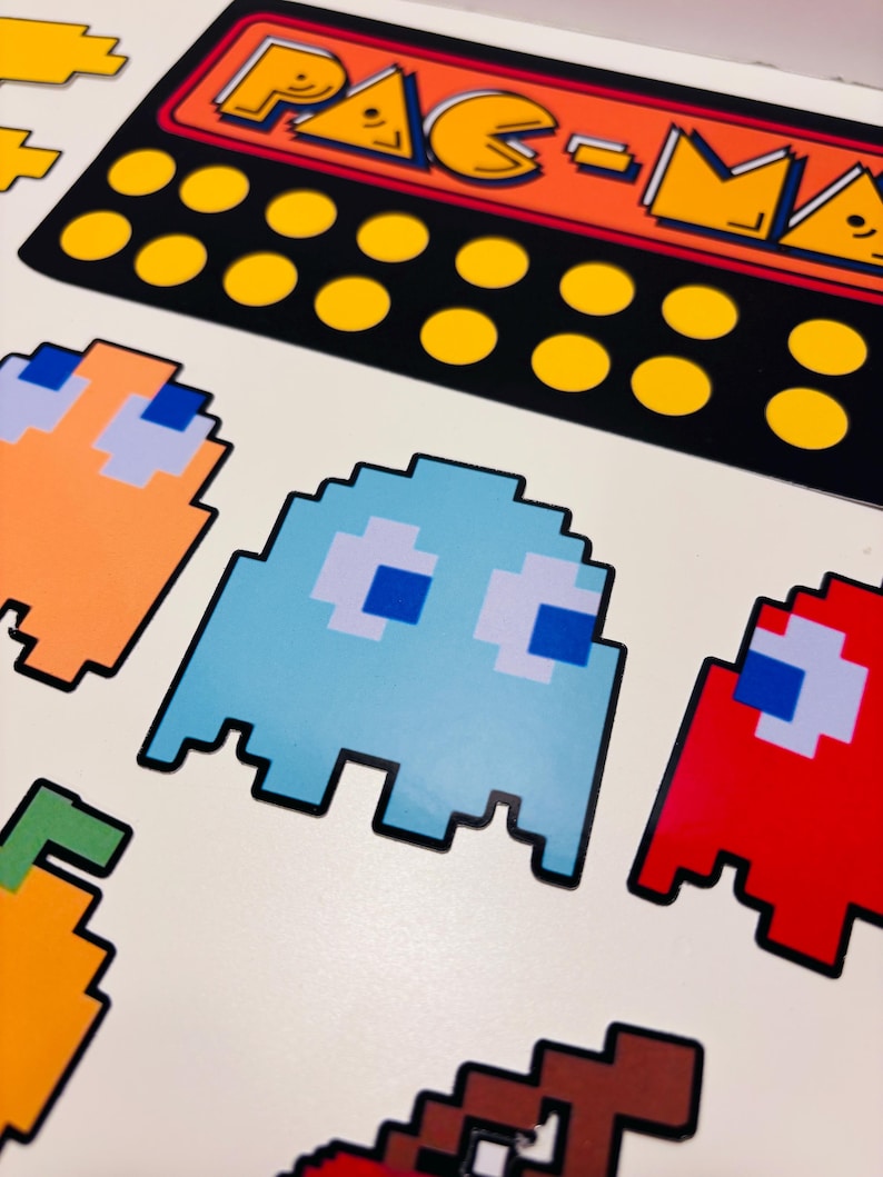 以下が含まれることがあります： ピクセル化されたパックマンのキャラクターとゲーム要素が描かれたステッカーシート。赤、オレンジ、水色のゴースト、黄色のドット、そしてオレンジと黄色の文字でPAC-MANと書かれた黒い長方形のロゴが含まれています。