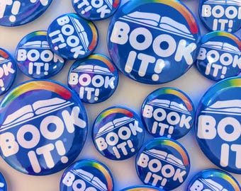 Könnte beinhalten: Eine Sammlung runder blauer Anstecknadeln mit dem weißen Text BOOK IT! und einem stilisierten weißen Symbol eines offenen Buches mit einem regenbogenfarbenen Bogen darüber. Die Anstecknadeln variieren in der Größe und sind zufällig angeordnet.
