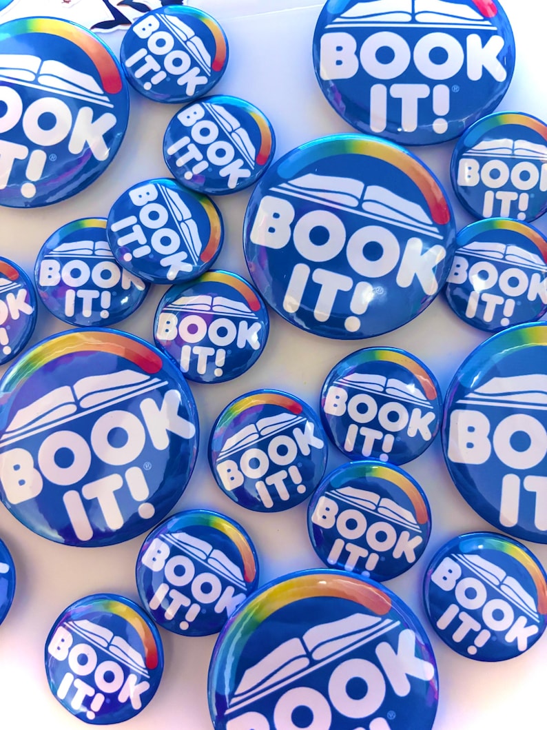 K&ouml;nnte beinhalten: Eine Sammlung runder blauer Ansteckbuttons mit dem wei&szlig;en Text BOOK IT!, einer Grafik eines wei&szlig;en offenen Buches und einem regenbogenfarbenen Bogen dar&uuml;ber. Die Buttons haben unterschiedliche Gr&ouml;&szlig;en und sind verstreut angeordnet.