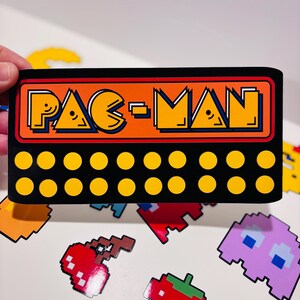 以下が含まれることがあります： 濃い青色の輪郭を持つオレンジ色のパックマンのロゴが描かれた長方形の黒いステッカー。ロゴの下には、それぞれ10個の黄色い円が並んだ2つの水平な列があります。ステッカーは、さまざまなピクセル化されたゲームアイコンの背景に対して表示されています。