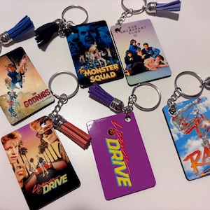 May include: Keychains featuring movie posters from the 1980s, including "The Goonies," "Monster Squad," "The Breakfast Club," "License to Drive," and "Rad." Each keychain has a metal ring, chain, and a tassel in various colors.