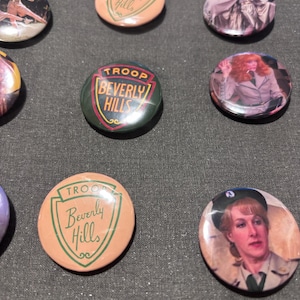 May include: A collection of round pinback buttons. Some buttons feature the text "TROOP Beverly Hills" in a shield design. Other buttons display images of characters. The buttons are in various colors.