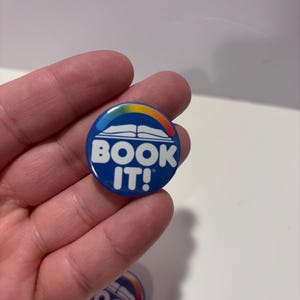 K&ouml;nnte beinhalten: Ein runder blauer Ansteckbutton mit einer wei&szlig;en Grafik eines offenen Buches, das von einem mehrfarbigen Regenbogenbogen gekr&ouml;nt wird. Der Text "BOOK IT!" ist in fetten wei&szlig;en Gro&szlig;buchstaben unter der Buchgrafik auf dem runden Button abgedruckt.