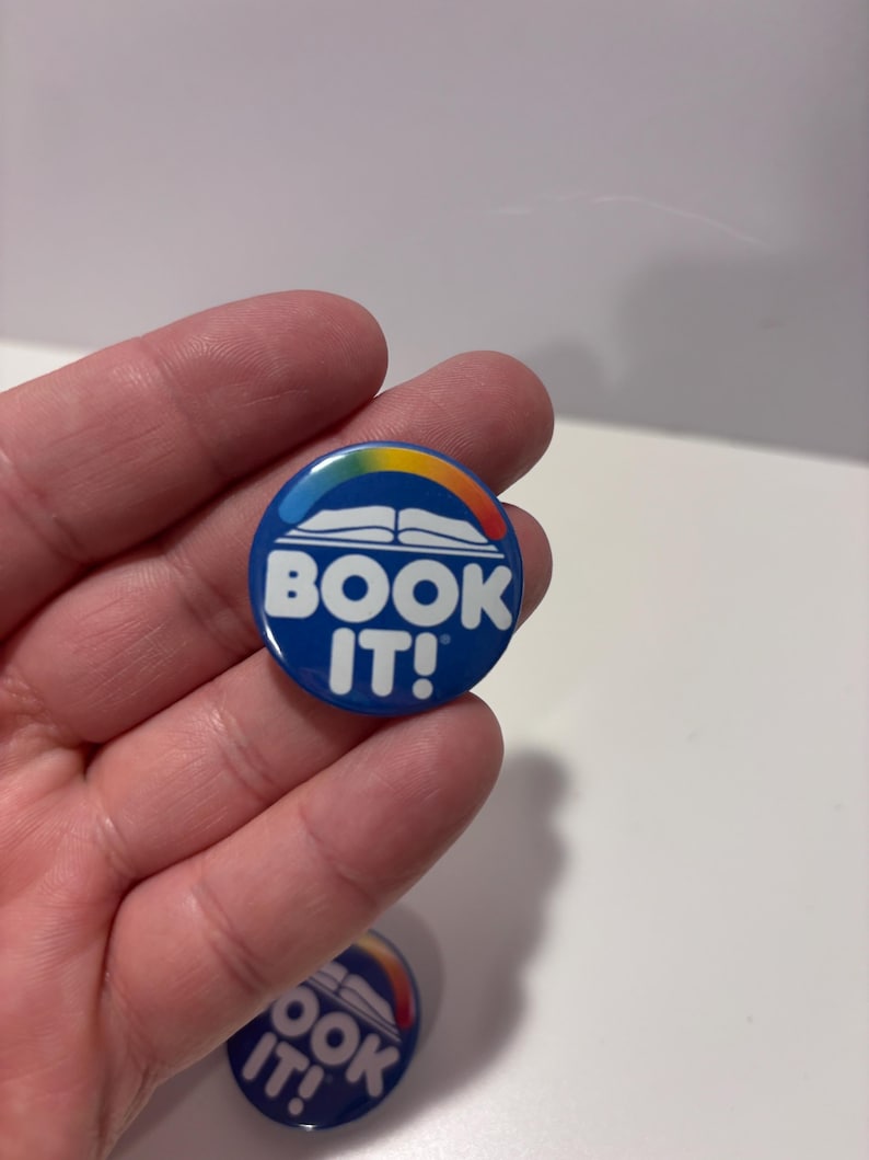 K&ouml;nnte beinhalten: Ein runder blauer Ansteckbutton mit einer wei&szlig;en Grafik eines offenen Buches, das von einem mehrfarbigen Regenbogenbogen gekr&ouml;nt wird. Der Text "BOOK IT!" ist in fetten wei&szlig;en Gro&szlig;buchstaben unter der Buchgrafik auf dem runden Button abgedruckt.