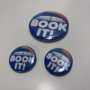 May include: Three blue round buttons with the text "BOOK IT!" in white, arched above an open book graphic and a rainbow. The buttons are on a white surface.