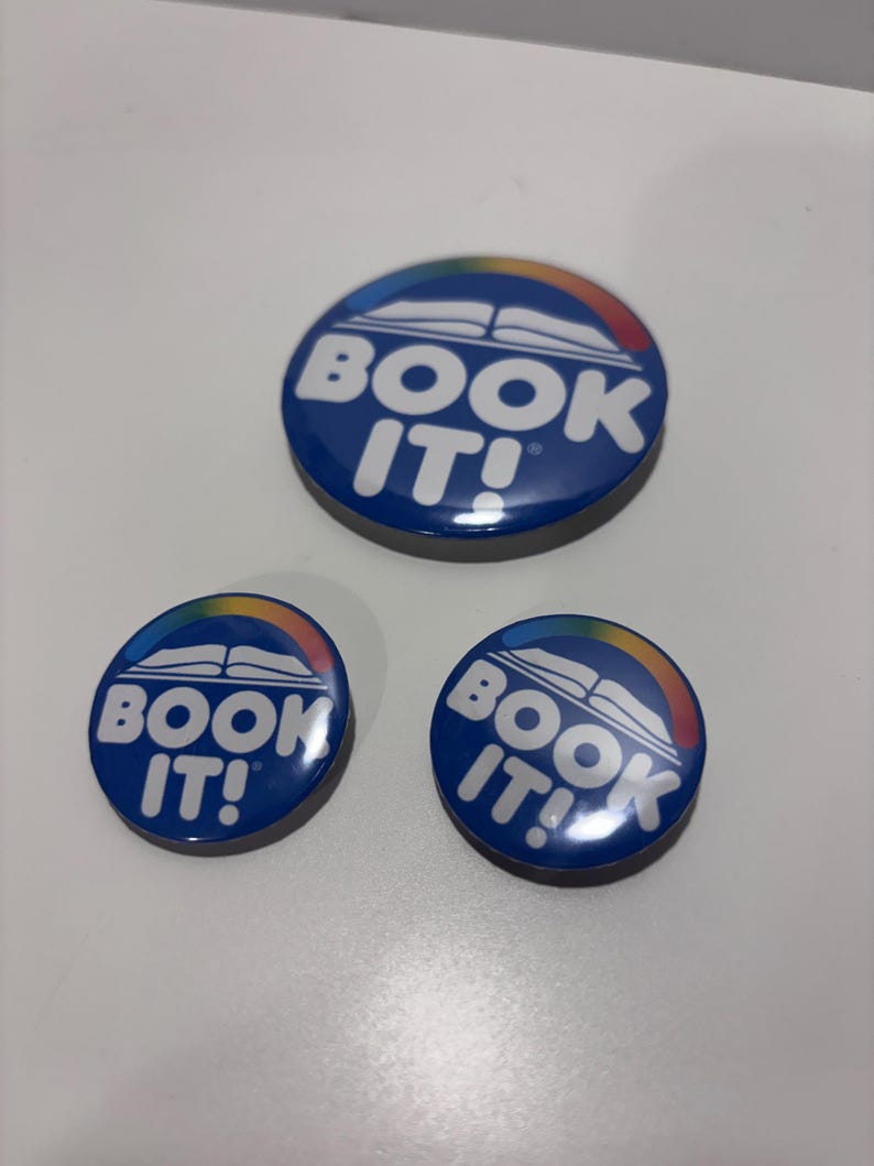 K&ouml;nnte beinhalten: Drei runde Ansteckbuttons in verschiedenen Gr&ouml;&szlig;en mit dunkelblauem Hintergrund und einer wei&szlig;en Grafik eines offenen Buches, das von einem mehrfarbigen Regenbogenbogen gekr&ouml;nt wird. Der Text "BOOK IT!" steht in wei&szlig;en Gro&szlig;buchstaben darunter.