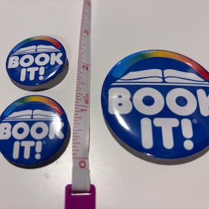 K&ouml;nnte beinhalten: Drei kreisf&ouml;rmige blaue Anstecknadeln mit dem wei&szlig;en Text BOOK IT!, einer wei&szlig;en Grafik eines offenen Buches und einem Regenbogenbogen dar&uuml;ber. Ein Ma&szlig;band liegt vertikal zwischen den Nadeln und zeigt, dass die gr&ouml;&szlig;te 7,6 cm gro&szlig; ist.