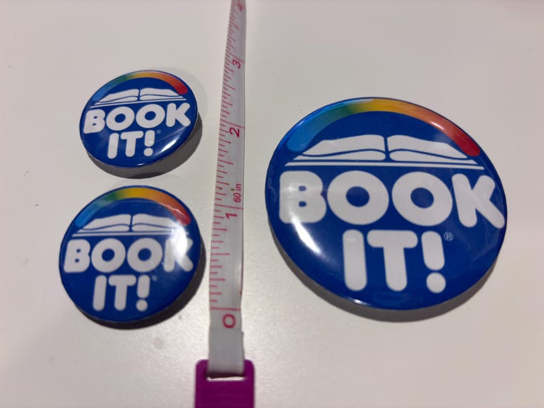 K&ouml;nnte beinhalten: Drei kreisf&ouml;rmige blaue Anstecknadeln mit dem wei&szlig;en Text BOOK IT!, einer wei&szlig;en Grafik eines offenen Buches und einem Regenbogenbogen dar&uuml;ber. Ein Ma&szlig;band liegt vertikal zwischen den Nadeln und zeigt, dass die gr&ouml;&szlig;te 7,6 cm gro&szlig; ist.
