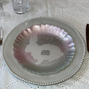 May include: A white ceramic plate with a scalloped edge, featuring a smaller iridescent plate with a bunny silhouette. The smaller plate has the text "Hand Made, Hand Wash Only, Made in Turkey".