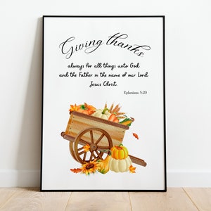 以下が含まれることがあります： 「Giving thanks」のテキストと聖書の詩が書かれた額入りのプリント。このアートワークは、カボチャ、カボチャ、トウモロコシでいっぱいの木製のカートが特徴です。カラーパレットには、オレンジ、黄色、茶色の色合いが含まれており、収穫のテーマを連想させます。