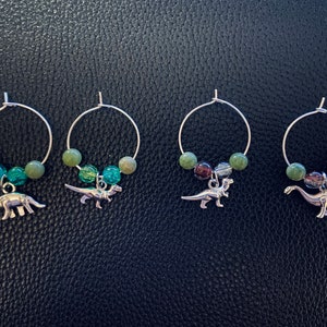 May include: Four silver wine charms with dinosaur pendants. Each charm has a silver hoop with green and blue beads. The dinosaur charms are different types, including a Brontosaurus and a T-Rex.