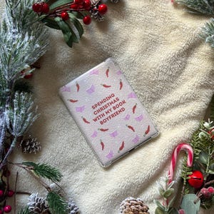 May include: A clear phone case with a light pink background, decorated with open book and chilli pepper motifs. The text "SPENDING CHRISTMAS WITH MY BOOK BOYFRIEND" is printed on the case. The case is surrounded by festive Christmas decorations.