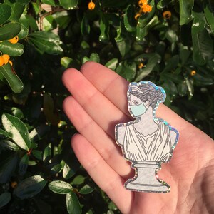 May include: A silver glitter sticker of a bust of a woman wearing a blue face mask. The woman is wearing a white toga-like garment. The sticker is on a hand.