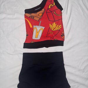 May include: A two-piece outfit featuring a red crop top and black shorts. The crop top has a black trim and is patterned with fast food graphics. The shorts are high-waisted and made of a textured black fabric.